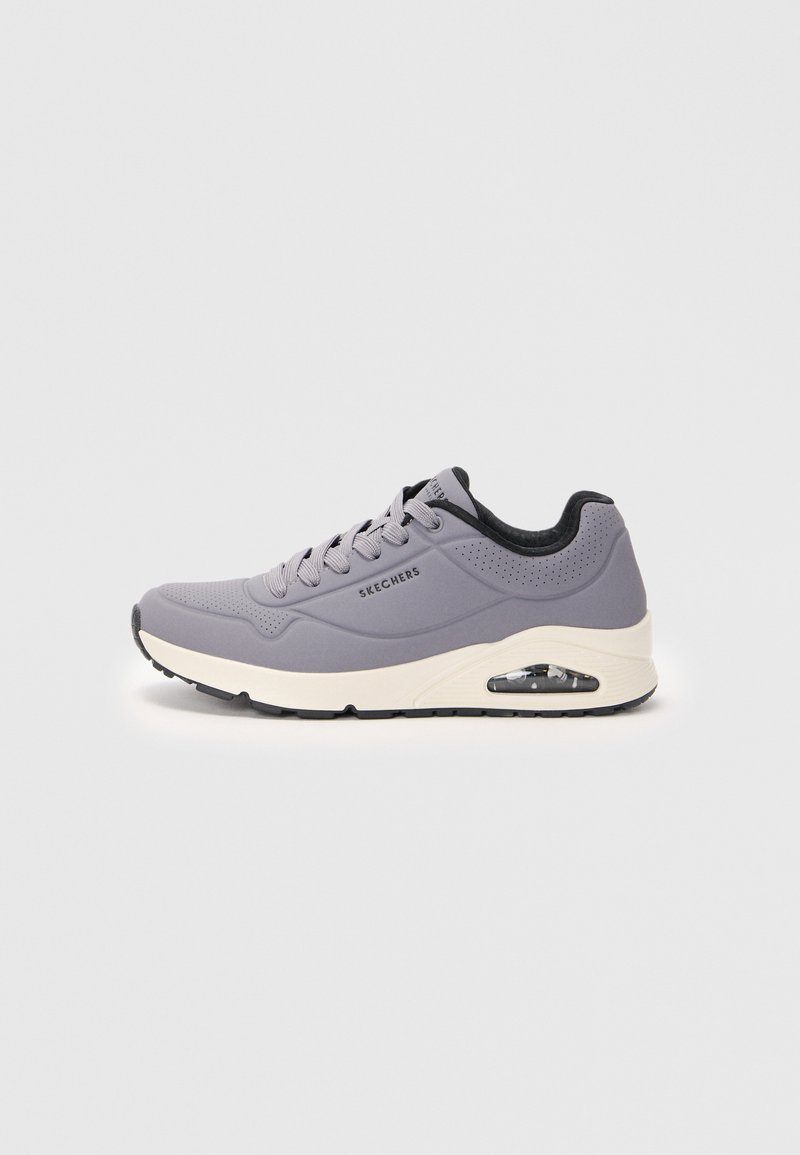 Harmaat Skechers-tennarit valkoisella pohjalla, mustalla pehmustetulla kauluksella, rei'itetty päällinen, näkyvä ilmatyyny ja nauhoilla kiristettävä etuosa tasaisella taustalla.
