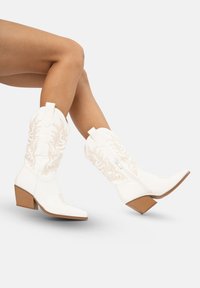 Botas de vaquero blancas con puntera cuadrada y un tacón bloque de altura media, con bordado beige y un detalle de cremallera lateral.