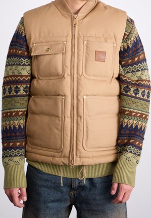 Person trägt eine hellbraune gesteppte Weste mit vier vorderen Taschen über einem grün-blau-braun gemusterten Strickpullover und blauen Jeans.