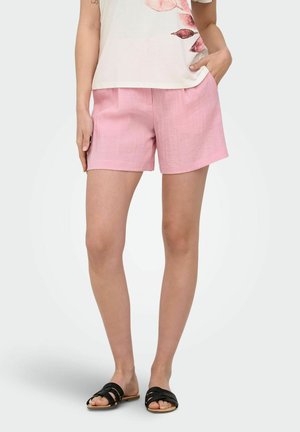 Femme portant un short rose clair, un haut blanc avec un imprimé de feuilles roses et des sandales noires à enfiler, debout, une main dans la poche.