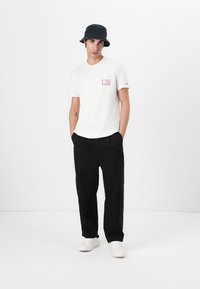 T-shirt en coton blanc avec un petit graphisme coloré sur la poitrine, associé à un pantalon noir ample et des baskets blanches, surmonté d'un chapeau seau bleu marine.