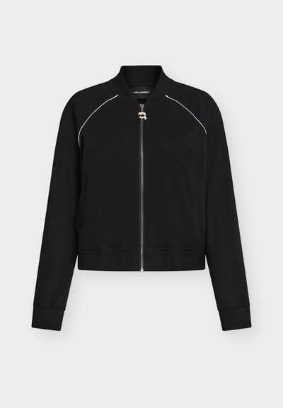 Veste bomber noire zippée avec un passepoil blanc aux épaules, col et poignets côtelés, et une petite tirette de fermeture éclair argentée en forme de visage.