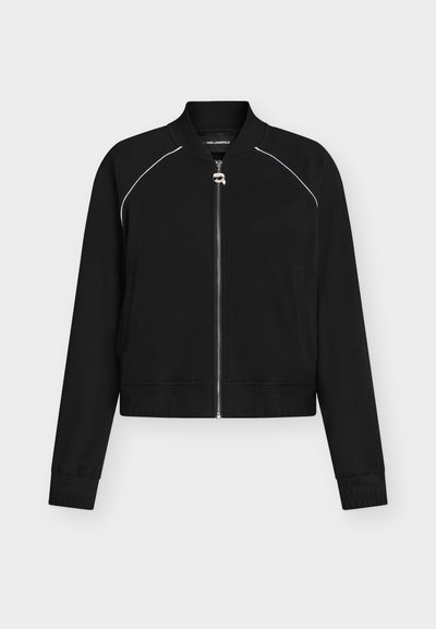 Veste bomber noire zippée avec un passepoil blanc aux épaules, col et poignets côtelés, et une petite tirette de fermeture éclair argentée en forme de visage.