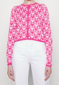 Różowo-biały skrócony kardigan z motywem serca, okrągłym dekoltem i zapięciem na guziki. Wykonany z miękkiego, dzianinowego materiału.