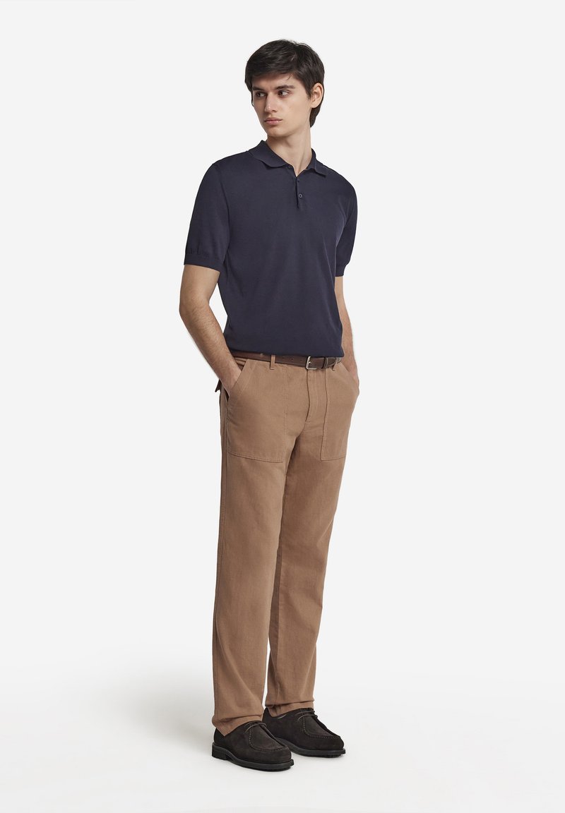 Marinblå pikétröja med korta ärmar och treknappsstängning, kombinerad med ljusbruna chinos med framfickor och ett brunt bälte.