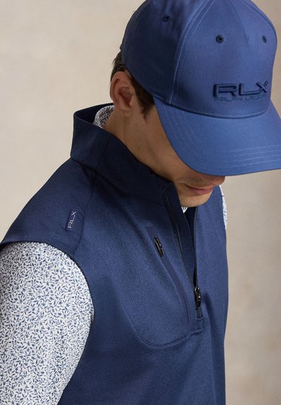 Homme portant une casquette bleu RLX et un gilet zippé bleu assorti par-dessus une chemise blanche avec des motifs de feuilles bleues, regardant vers le bas.