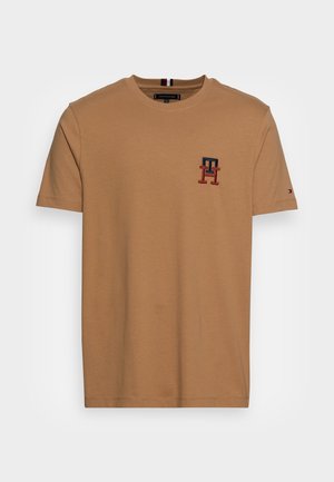 Camiseta de manga corta y cuello redondo en algodón marrón claro, con un pequeño logo bordado en azul marino y rojo en el lado izquierdo del pecho.