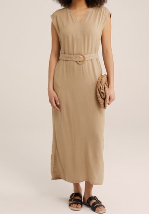 Kvinde iført en beige midi-kjole med bælte, der holder en flettet clutch og har sorte sandaler med gyldne spænder mod en neutral baggrund.