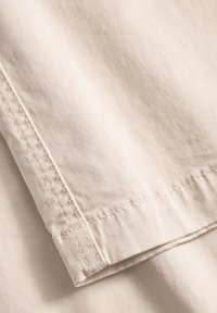 Heller beiger Stoff mit einer glatten Textur, der eine gefaltete Kante und sichtbare Nähte aufweist. Das Material wirkt robust und schlicht.