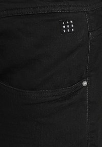 Pantaloni in denim neri con tasca laterale, dettagli cuciti e un piccolo etichetta quadrata con "DNM WORKRS" in lettere bianche.