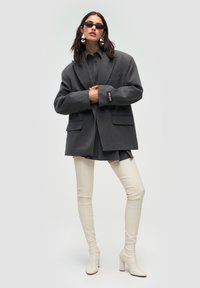 Donna che indossa un blazer over-size grigio scuro, una gonna corta coordinata, stivali alti fino alla coscia beige, occhiali da sole neri e grandi orecchini d'argento, in posa con sicurezza.