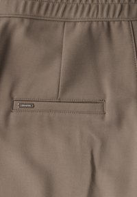 Bruna tygshorts med en bakficka med en metallisk varumärkesetikett och subtila sömmar längs midjan. Slät textur.