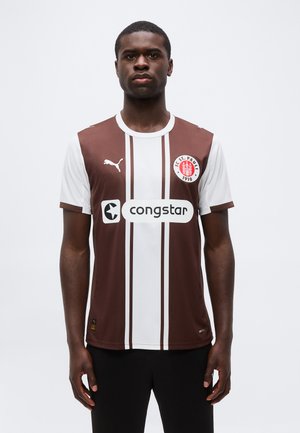 Mann iført brun og hvit FC St. Pauli fotballdrakt med "congstar" sponsorlogo, står mot en ensfarget lys bakgrunn.