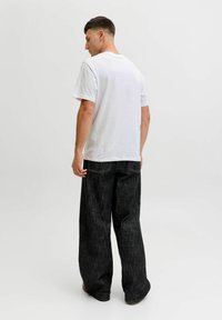 Hombre de espaldas con una camiseta blanca de manga corta y pantalones de mezclilla negros holgados, frente a un fondo claro y liso.