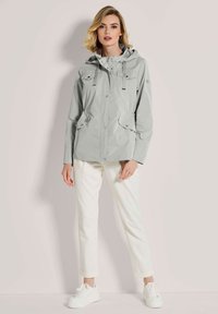 Hellgraue wasserdichte Jacke mit Kapuze, Knopfverschlüssen, Fronttaschen und Kordelzügen. Getragen über weißen Hosen und Turnschuhen.