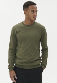 Olivově zelená mikina s dlouhými rukávy a kulatým výstřihem, s žebrovanými manžetami a lemem, vyrobená z hladkého materiálu, nošená přes černé kalhoty.