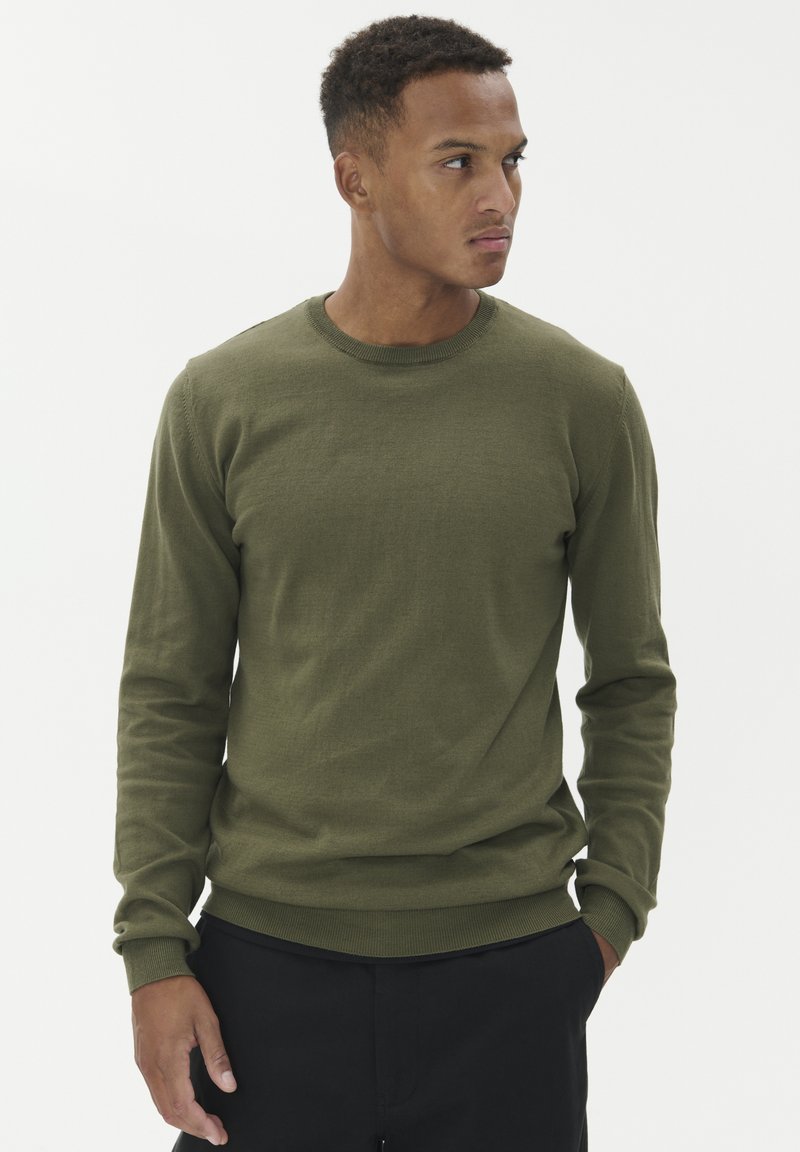 Olivově zelená mikina s dlouhými rukávy a kulatým výstřihem, s žebrovanými manžetami a lemem, vyrobená z hladkého materiálu, nošená přes černé kalhoty.