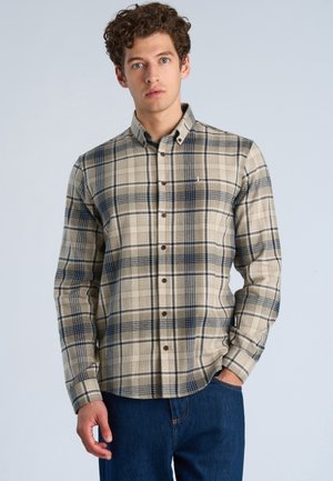 Junger Mann mit lockigem Haar, der ein beiges und marineblau kariertes Hemd und blaue Jeans trägt, steht mit einer Hand in der Tasche vor einem schlichten Hintergrund.