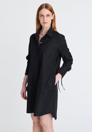 Frau mit glatten roten Haaren trägt ein langärmliges schwarzes Hemdblusenkleid mit Kragen, eine Hand in der Tasche, steht vor einem schlichten weißen Hintergrund.