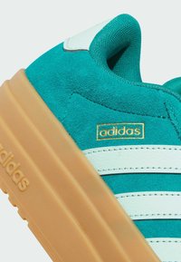 Baskets Adidas en daim bleu sarcelle et cuir blanc avec semelle en gomme, logo Adidas doré et bandes blanches cousues sur le côté.