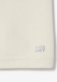 Gros plan d'un tissu beige avec un petit texte argenté "ULTRA DRY" cousu près du bord inférieur.