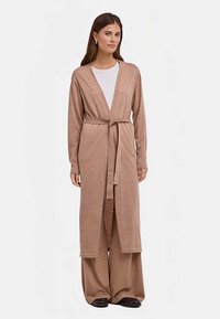 Cardigan long beige avec ceinture à nouer, conception à ouverture devant, texture douce et manches longues, associé à un pantalon beige à jambes larges.
