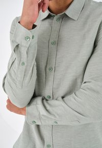 Uomo che indossa una camicia a maniche lunghe verde chiaro, con le braccia incrociate e una mano che tocca il viso.