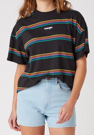 Frau trägt ein schwarzes Wrangler T-Shirt mit bunten horizontalen Streifen und hellblauen Denim-Shorts, gezeigt von den Schultern bis zur Mitte der Oberschenkel.