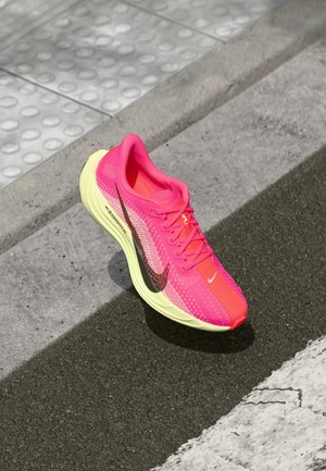 Helles pinkfarbenes Laufschuh mit neon-gelber Sohle und schwarzem Swoosh-Logo, auf Asphalt in der Nähe eines Bürgersteigrands platziert.