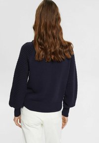 Pull en maille bleu marine avec manches bouffantes texturées, ourlet côtelé et col rond. Porté avec un pantalon clair.