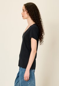 Moteris su ilgais garbanotais plaukais, dėvinti juodą V formos kaklo trumpomis rankovėmis palaidinę ir mėlynas džinsus, stovinti profiliu priešais paprastą smėlio spalvos foną.