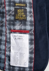 Tamsus švarkas su margintu pamušalu, su etikete "OPPOSUITS" ir juokingu valgymo gidu, apibrežtu juodu ant balto audinio.