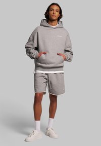 Felpa grigia con grande cappuccio e tasche laterali, abbinata a short grigi coordinati con rifiniture bianche e cordino. Indossata con sneaker bianche.