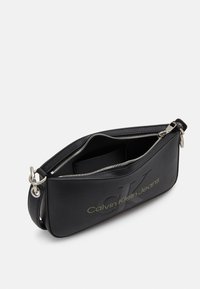 Crna kožna torbica s srebrnim zatvaračem. Sadrži urezani "CK" logo i "Calvin Klein Jeans" u zelenoj boji. Uključuje odvojivi remen.