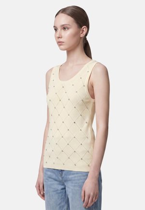 Giovane donna con capelli castano chiaro raccolti indietro, indossa un top giallo pallido senza maniche con motivo a rombi e jeans azzurri chiari.