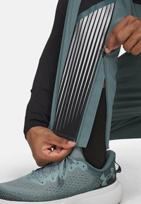 Sportieve broek met een ritsopening, teal met zwarte en zilveren streepaccenten, gecombineerd met grijze mesh sneakers. Close-up van een hand die de rits aanpast.