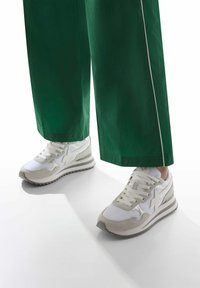 Baskets de sport avec une tige blanche, accents en daim gris clair, semelle texturée et lacets. Associées à un pantalon large vert avec des bordures blanches.