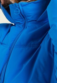 Niebieska kurtka puchowa z wysokim kołnierzem, prezentująca gładką teksturę i frontowy zamek błyskawiczny. Charakteryzuje się minimalnym przeszyciem oraz subtelną kieszenią na piersi.