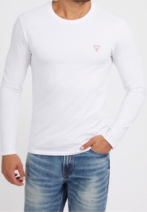 Mężczyzna w dopasowanej białej koszuli z długim rękawem, z małym czerwonym trójkątnym logo na klatce piersiowej, noszący wyblakłe niebieskie dżinsy, z ręką w kieszeni.