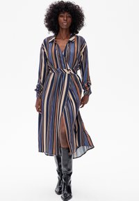Sukienka w paski w niebieskim, brązowym i kremowym kolorze, z zapinanym w talii, kołnierzykiem i długimi rękawami; posiada wysoki rozporek i zwiewny materiał.