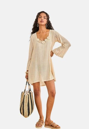 Mujer con vestido beige de manga larga con escote floral, sosteniendo un bolso tejido a rayas y llevando sandalias con tiras trenzadas.