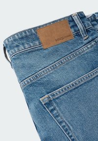 Modré džínové kalhoty z denimu s hnědým koženým štítkem označeným "MNG DENIM." Obsahují detaily stehu a design s pěti kapsami. Hladká textura.