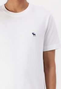 Maglietta bianca in cotone con colletto rotondo e maniche corte. Presenta un piccolo logo di un alce ricamato di colore blu sulla parte sinistra del petto.