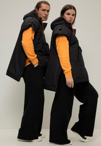 Gilet a vento neri con cappuccio, dettagli arancioni, indossati sopra felpe gialle. Modelli in pantaloni larghi neri e sneaker bianche.