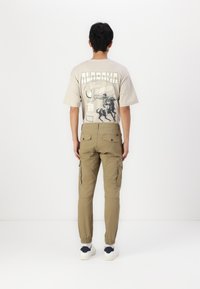 Beige grafisch T-shirt met "Alabama State Rodeo" print; tan cargobroek met zijzakken en elastische manchetten; witte sneakers met marineblauwe accenten.