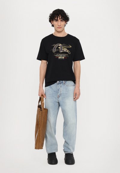 Černé tričko s grafickým designem, světle modré džíny a hnědá taška. Uzavřené černé boty. Jednoduchý, neformální outfit.