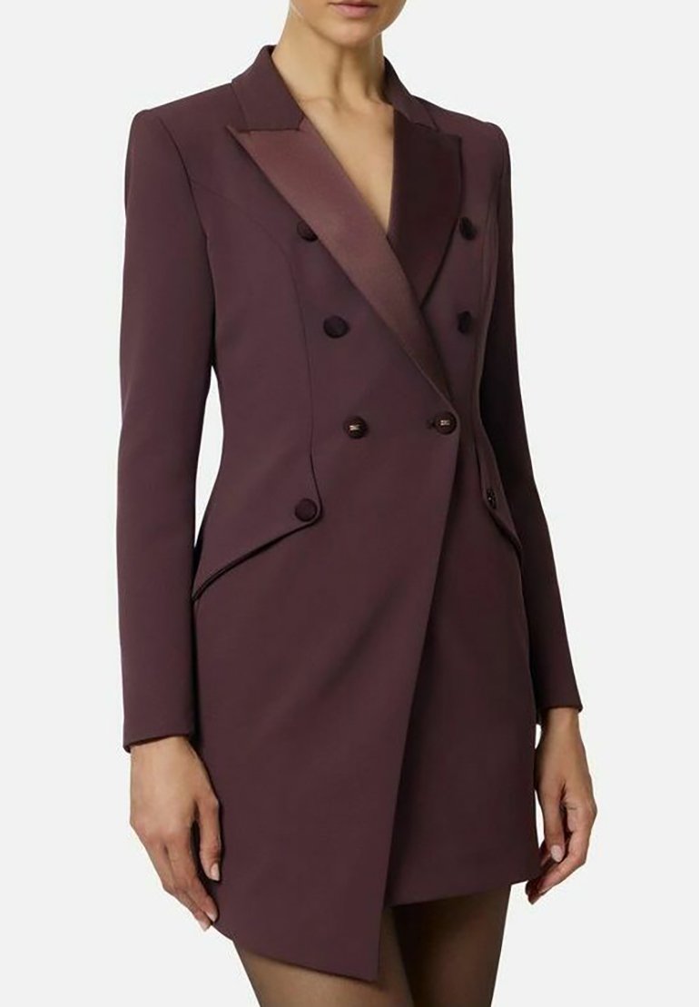 Abito blazer doppiopetto in bordeaux profondo, con spalle imbottite, dettagli in raso e revers a lancia. Tessuto liscio, vestibilità sagomata.