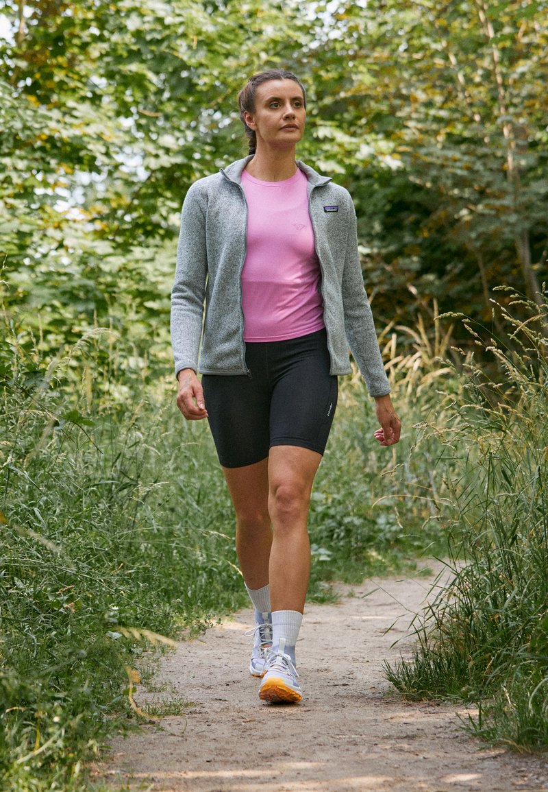 Graue Fleece-Jacke mit Reißverschluss über einem pinken T-Shirt und schwarzen Shorts, mit weißen Knöchelsocken und grauen Laufschuhen, die auf einem Weg gehen.