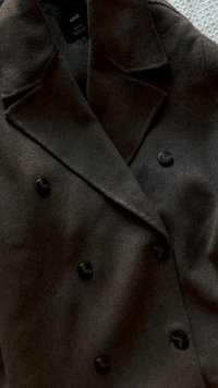 Cappotto doppiopetto in tessuto marrone scuro, con una morbida texture e grandi bottoni neri. Presenta un colletto a punta e un design sartoriale.