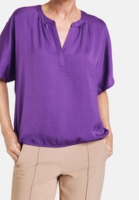 Lila kortärmad blus med v-ringning, tillverkad av slät tyg. Bärs över ljusbeiga byxor, med subtila samlingar vid halsen.